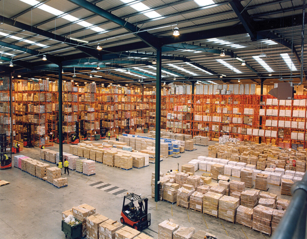 Wide warehouse interior with tall pallet racks, stacked cartons and forklifts.